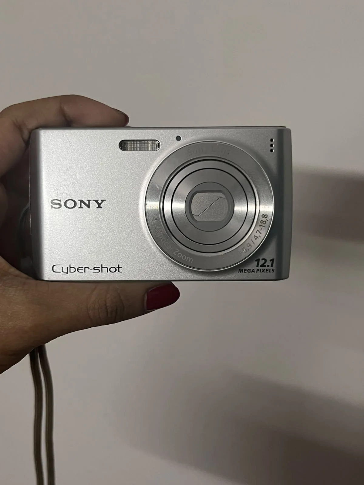 A hand holding a small silver Sony Cyber-shot point-and-shoot camera against a white wall.