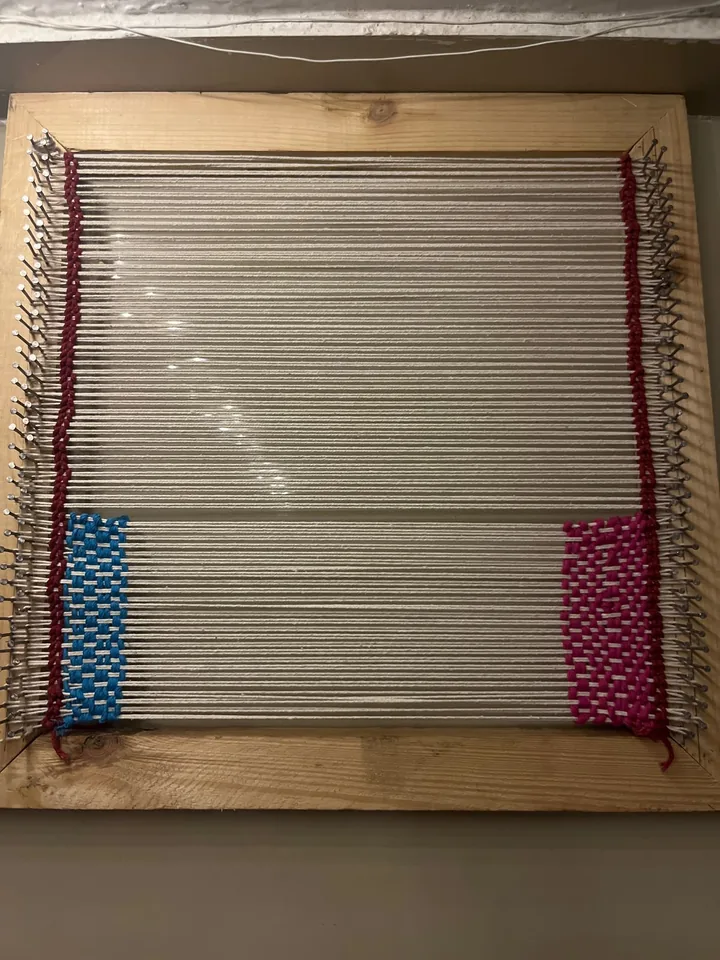 A wooden frame loom warped with cream thread, with the first blue and pink sections woven in.