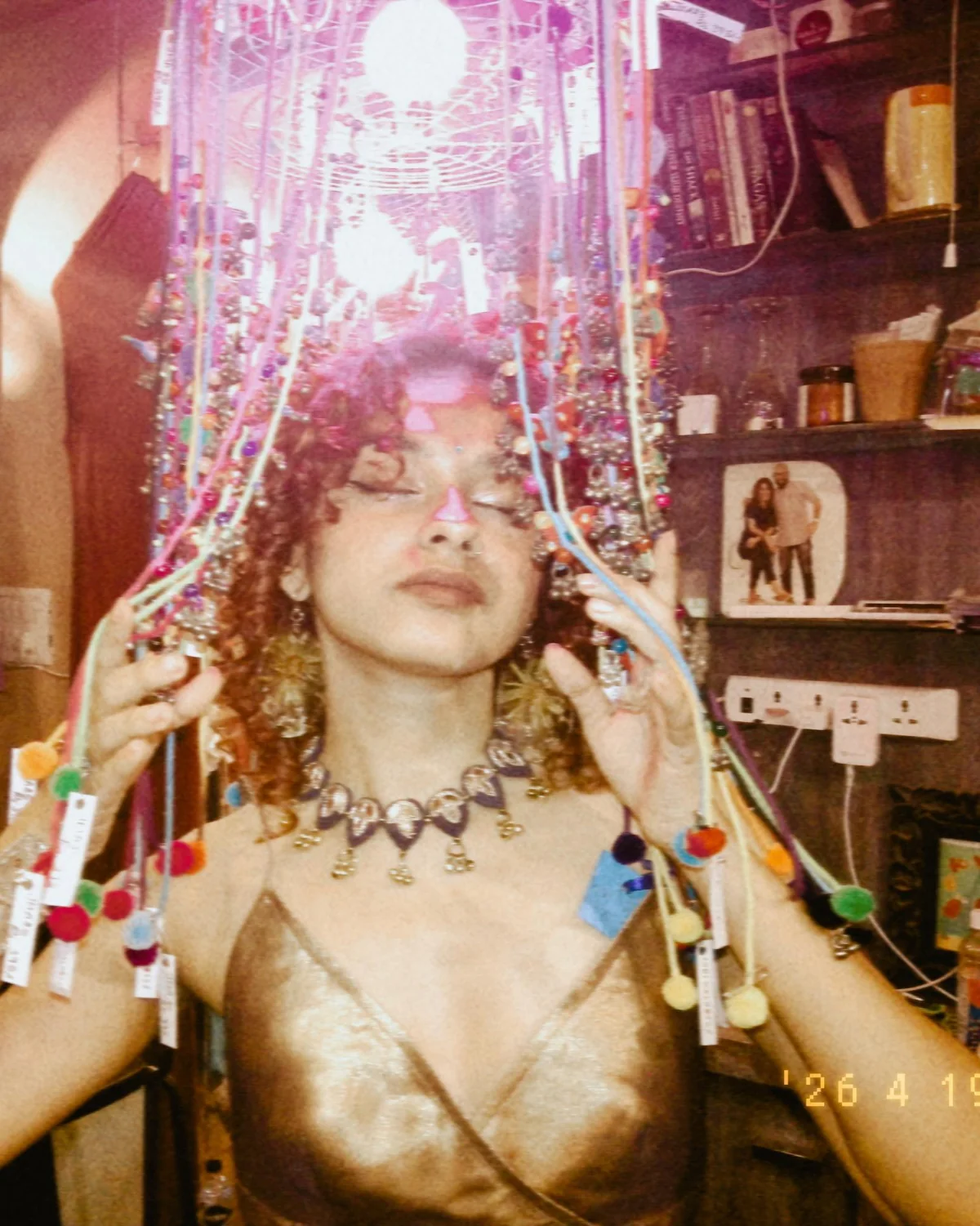 Shreshta with eyes closed under a canopy of hanging beaded strings and pom-poms, wearing a bronze satin top and a silver choker, lit pink.