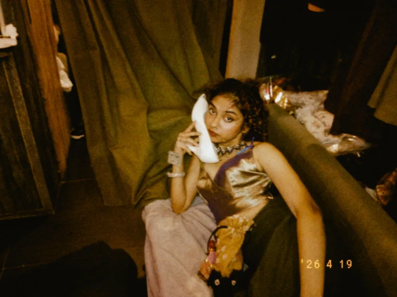 Shreshta sitting against olive curtains, holding a white heeled shoe to her ear like a telephone, in a bronze satin dress.