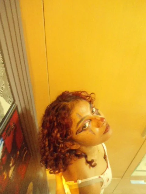 Shreshta lying on a yellow floor with red curls spread out, looking up at the camera with graphic eye makeup.