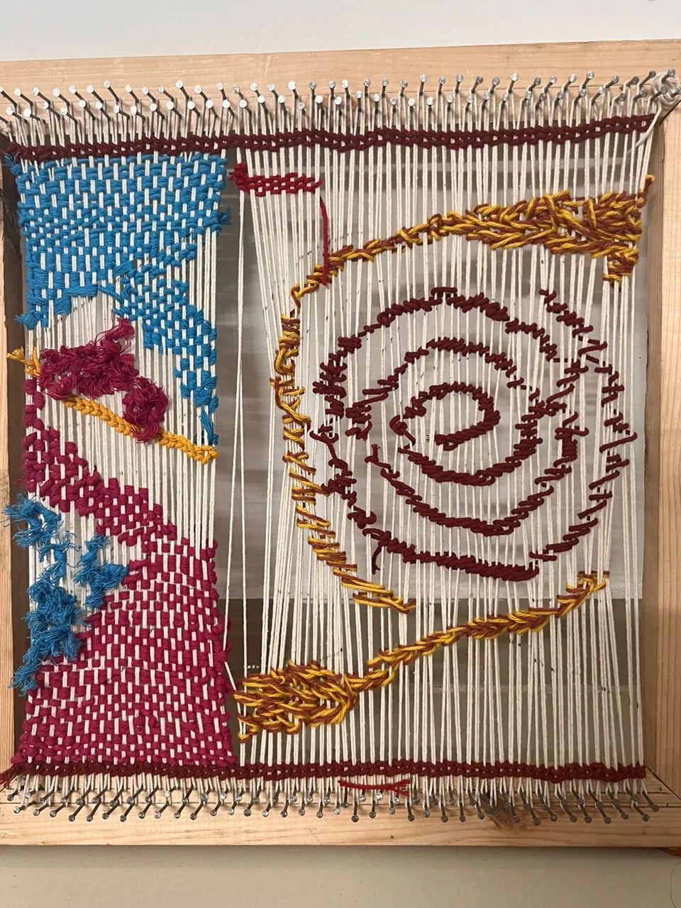 The final woven textile on a wooden frame loom: a crimson spiral at the centre, with blue, magenta and gold yarn radiating around it on a cream warp.